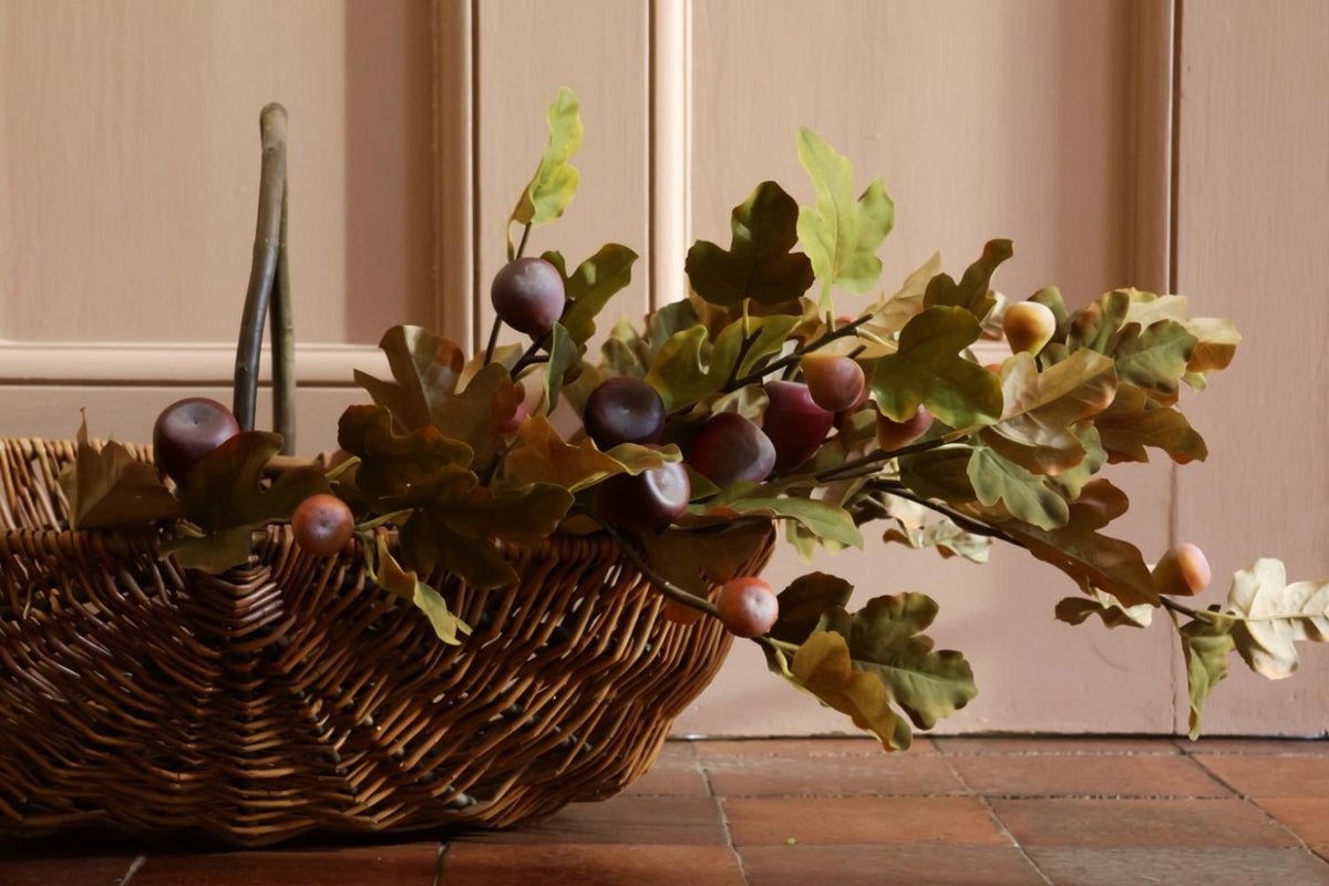 woven basket of artificial branches with fig and earthy toned leaves on a red tiled floor with pink door behind 