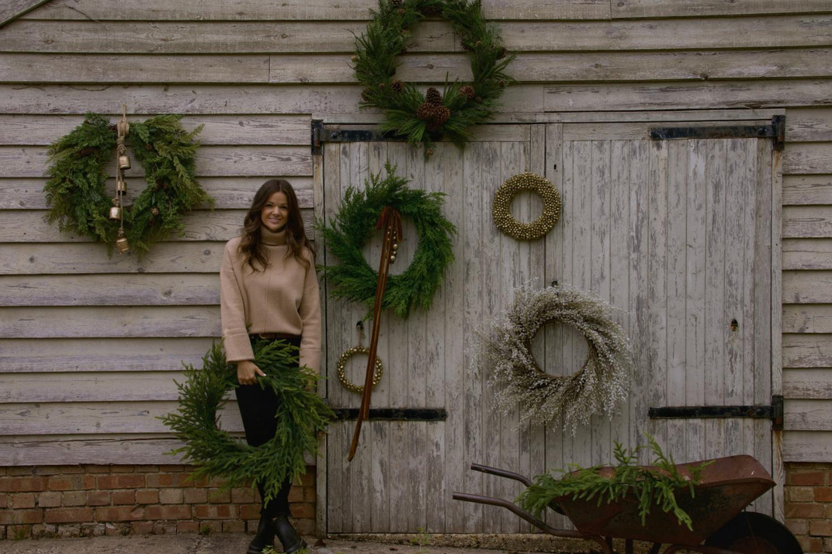 Artificial christmas wreaths positioned on a grey barn with lady holding a green wreath 