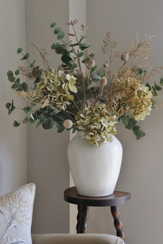 White vase with a bouquet of flowers on a wooden stool against a plain wall.