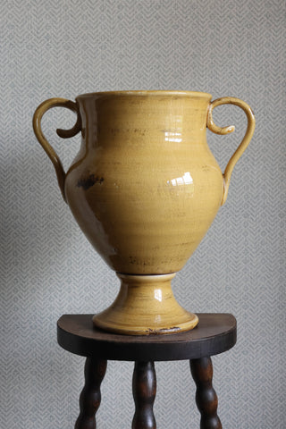 Yellow ceramic urn with handles on a wooden stand against a gray textured wall