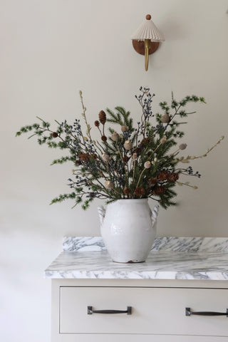 Decorative vase with greenery and pinecones on a marble surface with a neutral wall background.