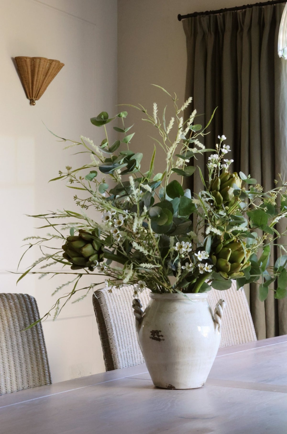 A large rustic white ceramic vase filled with a lush artificial green floral arrangement of eucalyptus, artichokes, and delicate wildflowers, set on a wooden dining table in a softly lit room.