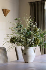 A large rustic white ceramic vase filled with a lush artificial green floral arrangement of eucalyptus, artichokes, and delicate wildflowers, set on a wooden dining table in a softly lit room.