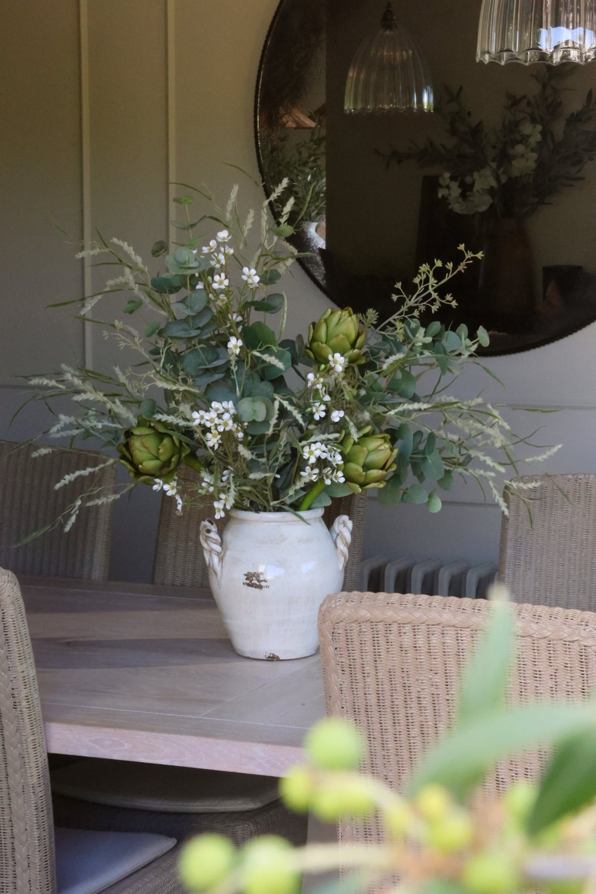A rustic white ceramic vase filled with a green and white floral arrangement of artichokes, eucalyptus, and small white waxflower, set on a dining table in a cozy room with a large round mirror in the background.