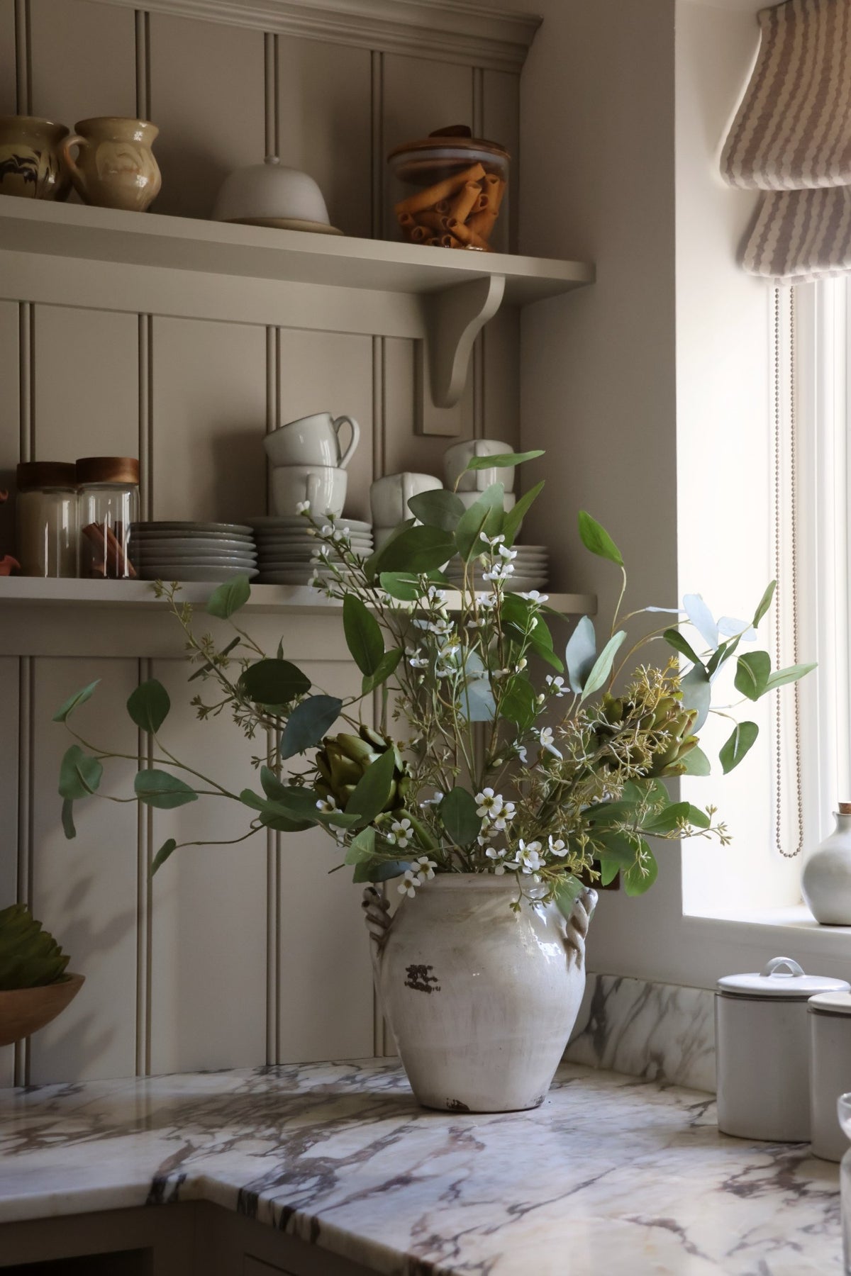 A rustic ceramic vase filled with artificial green eucalyptus, faux artichoke stems and white waxflower flowers on a marble kitchen counter, set against a paneled wall with shelves holding dishes.