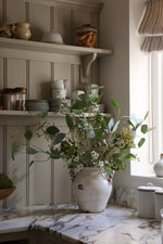 A rustic ceramic vase filled with artificial green eucalyptus, faux artichoke stems and white waxflower flowers on a marble kitchen counter, set against a paneled wall with shelves holding dishes.