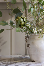 A rustic cream-colored ceramic vase filled with artificial faux green eucalyptus leaves, small white flowers, and a green artichoke, set on a marble countertop against a beige paneled wall.