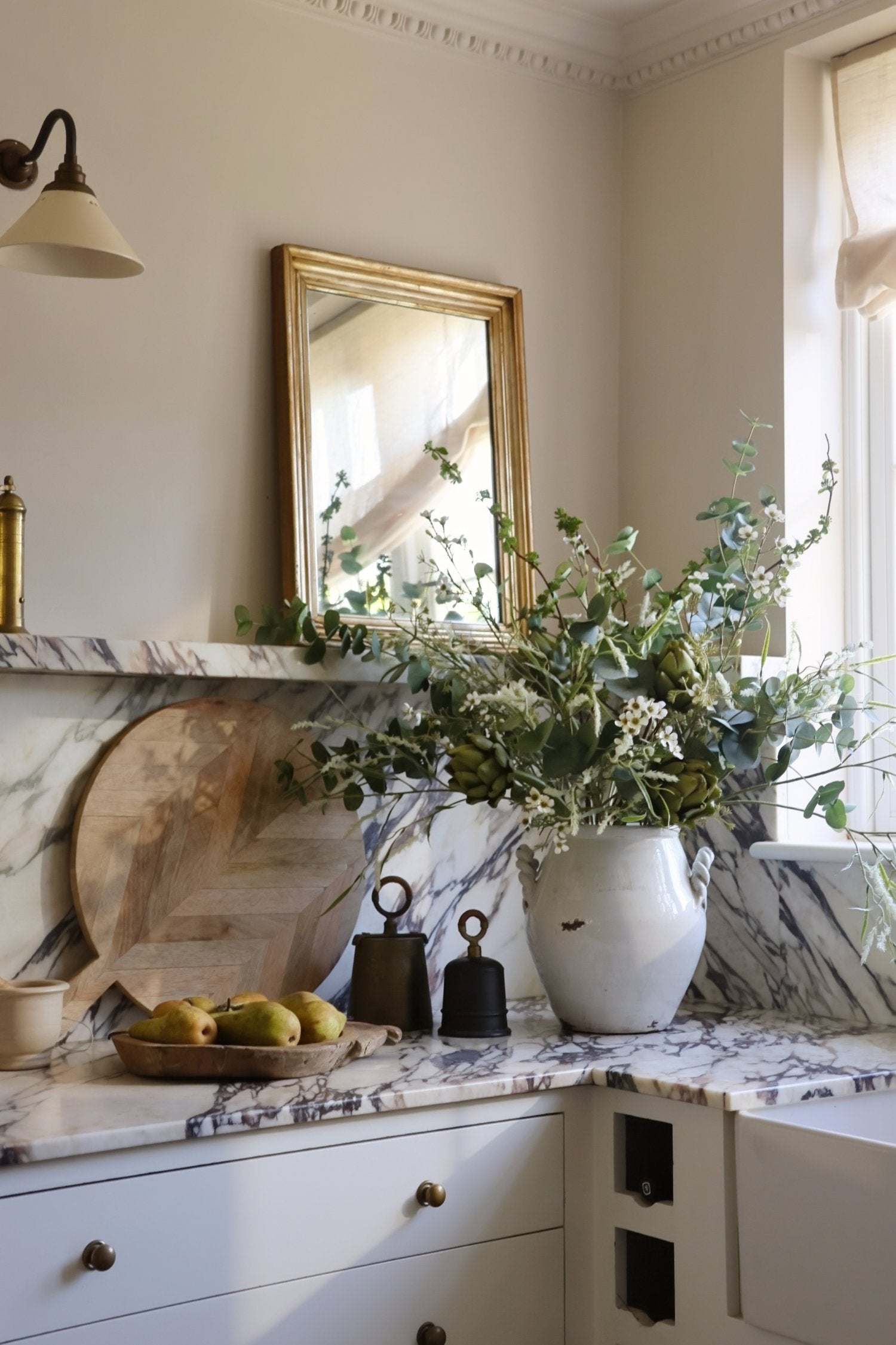 A styled kitchen corner featuring a white marble countertop with a matching marble backsplash, a large white ceramic vase filled with artificial foliage and artichoke stems.