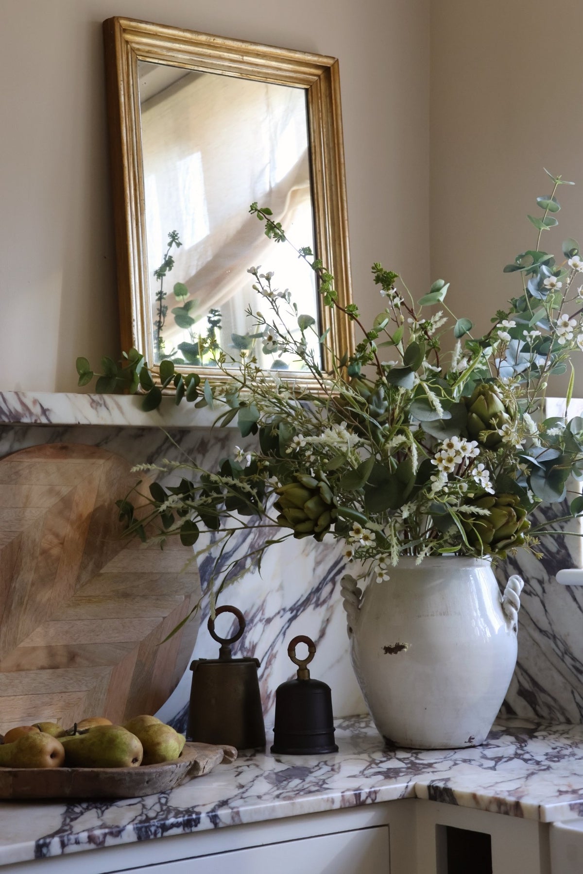 A rustic white ceramic vase filled with artificial green eucalyptus stems, waxflower and artichokes sits on a marble countertop next to two dark metal bells. 