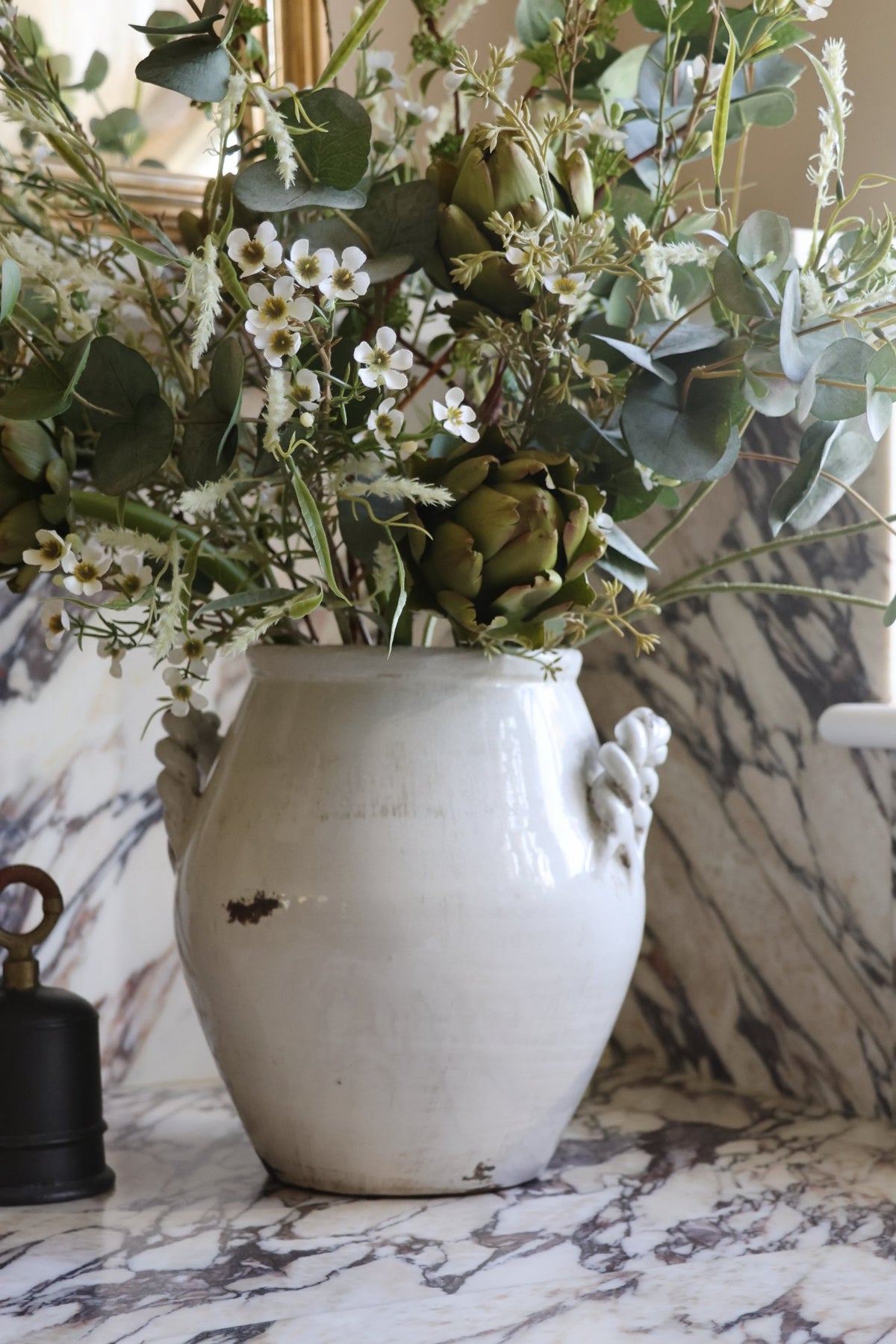 A rustic white ceramic vase with sculptural handles, filled with artificial green eucalyptus leaves, artichokes, and small white flowers, set on a white and gray marble surface.