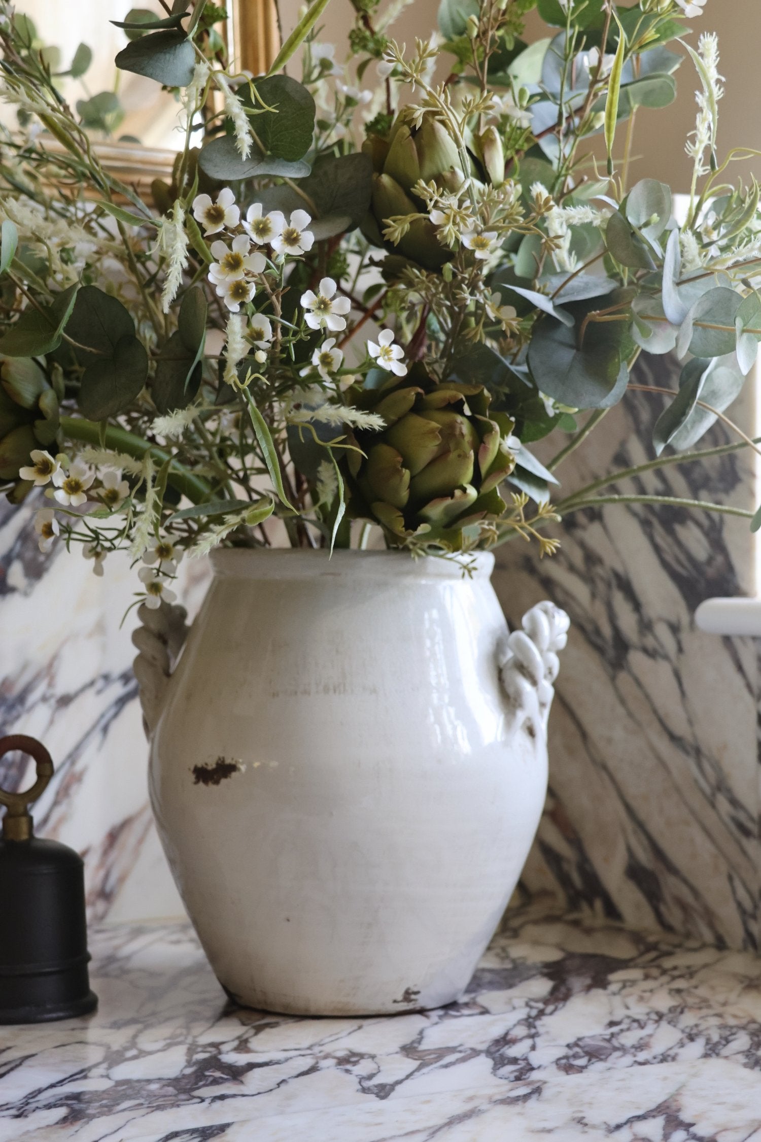 A rustic white ceramic vase with sculptural handles, filled with artificial green eucalyptus leaves, artichokes, and small white flowers, set on a white and gray marble surface.
