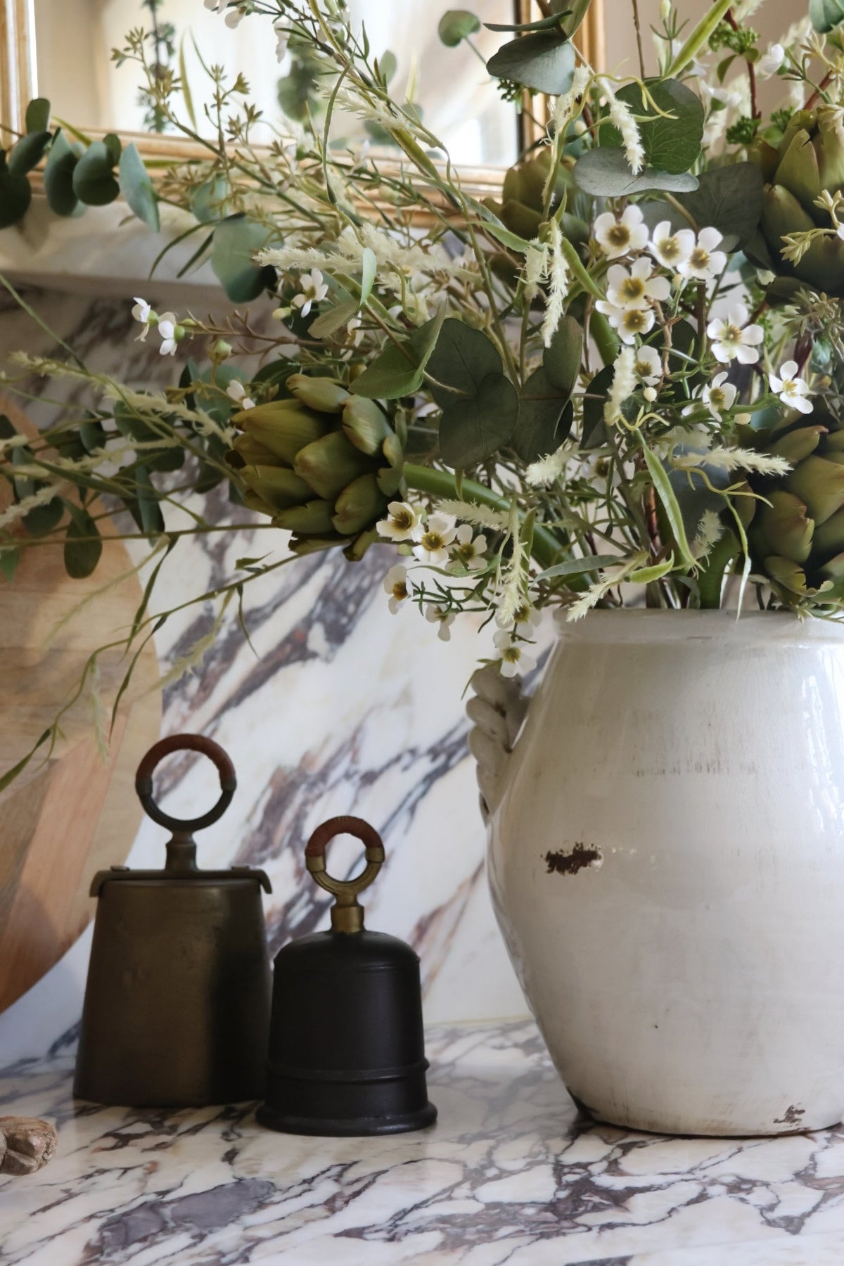 A rustic white ceramic vase filled with an artificial faux  floral arrangement featuring green eucalyptus, small white flowers, and artichoke stems, styled on a white and gray marble surface next to two vintage metal bells.
