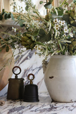 A rustic white ceramic vase filled with an artificial faux  floral arrangement featuring green eucalyptus, small white flowers, and artichoke stems, styled on a white and gray marble surface next to two vintage metal bells.