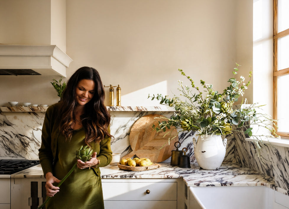 A woman with long dark hair, wearing an olive green dress, stands in a modern kitchen holding an artificial green artichoke. The kitchen features white marble countertops with black veining, a wooden cutting board, lemons, and a large white vase filled with artificial faux green foliage.