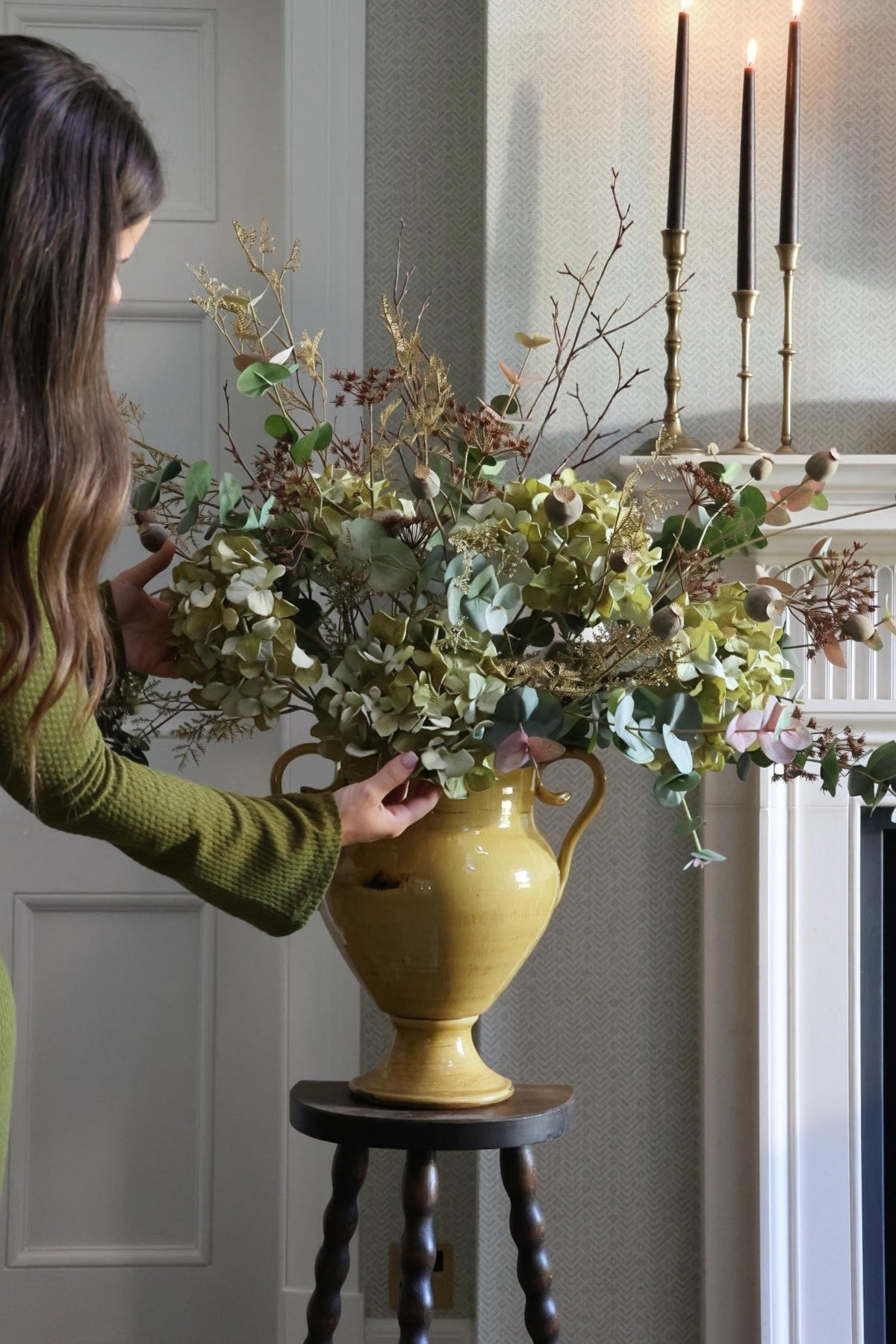Large artificial autumn floral arrangement in a mustard vase on a vintage stool featured dried green hydrangeas, eucalyptus and other autumn stems. 