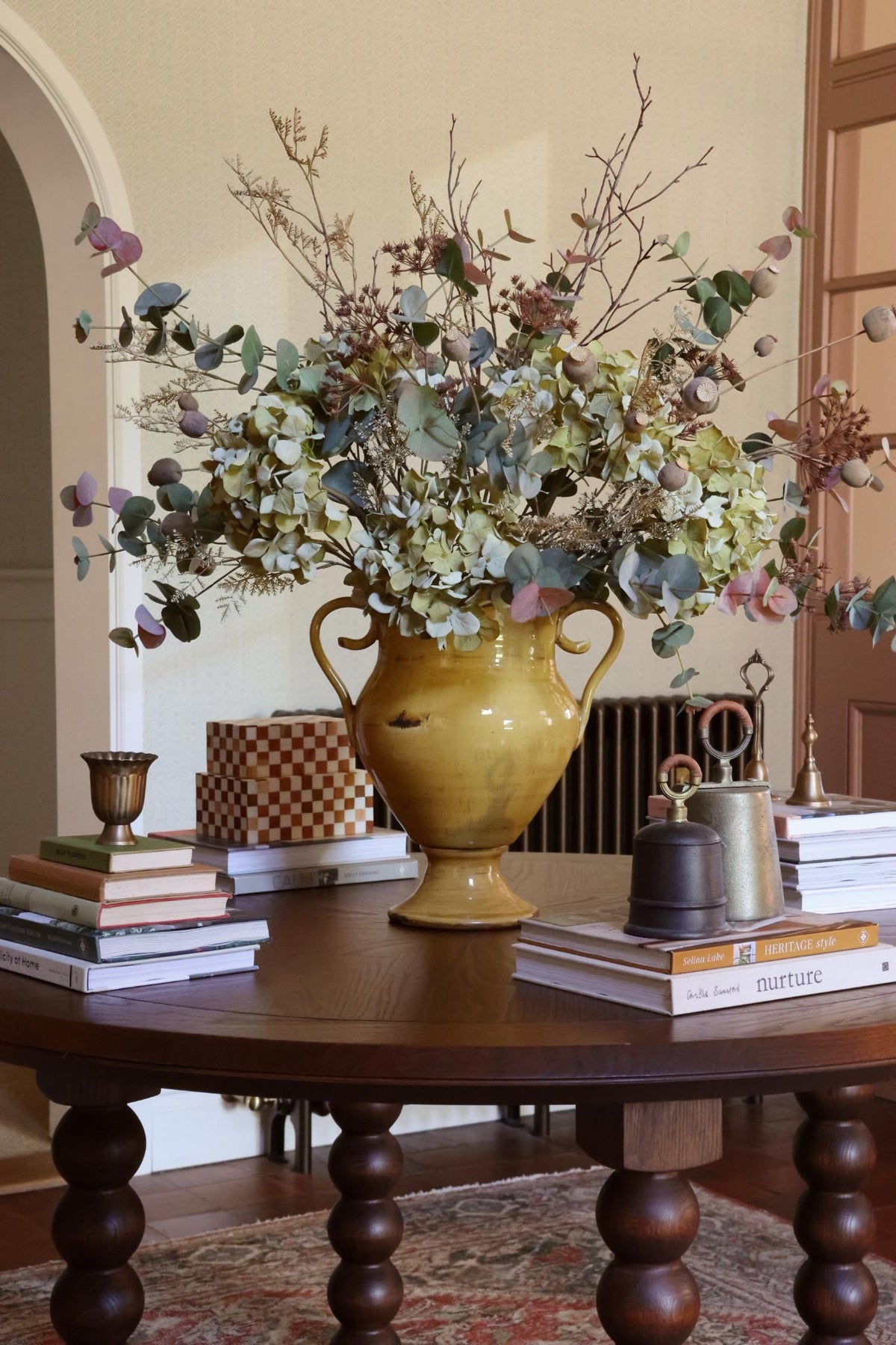 A large mustard yellow ceramic urn vase filled with artificial blush eucalyptus and other autumnal stems, displayed on a dark wood table in a warm, traditional room setting.