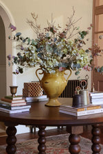 A large mustard yellow ceramic urn vase filled with artificial blush eucalyptus and other autumnal stems, displayed on a dark wood table in a warm, traditional room setting.