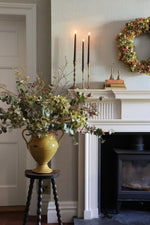 A large mustard ceramic vase filled with an autumnal artificial flower arrangement of dried green hydrangeas. The vase sits on a dark wooden stool in a living room setting next to a white fireplace mantel decorated with black taper candles, a floral wreath, and small books.