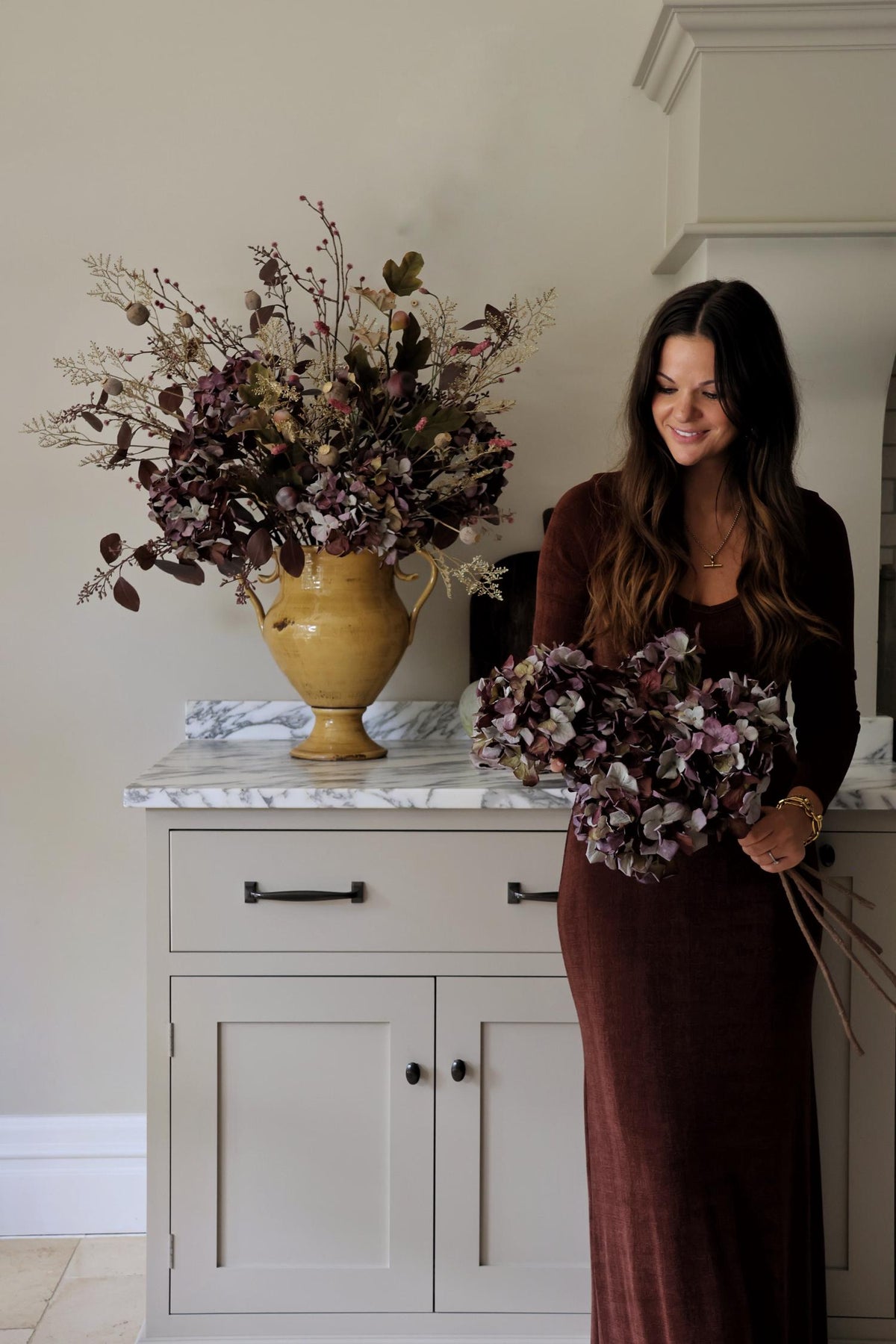 A woman in a long brown dress stands indoors beside a white cabinet, holding a bouquet of artificial dried burgundy hydrangeas. A large matching faux floral arrangement in a mustard vase sits on the cabinet counter.