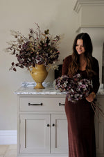 A woman in a long brown dress stands indoors beside a white cabinet, holding a bouquet of artificial dried burgundy hydrangeas. A large matching faux floral arrangement in a mustard vase sits on the cabinet counter.