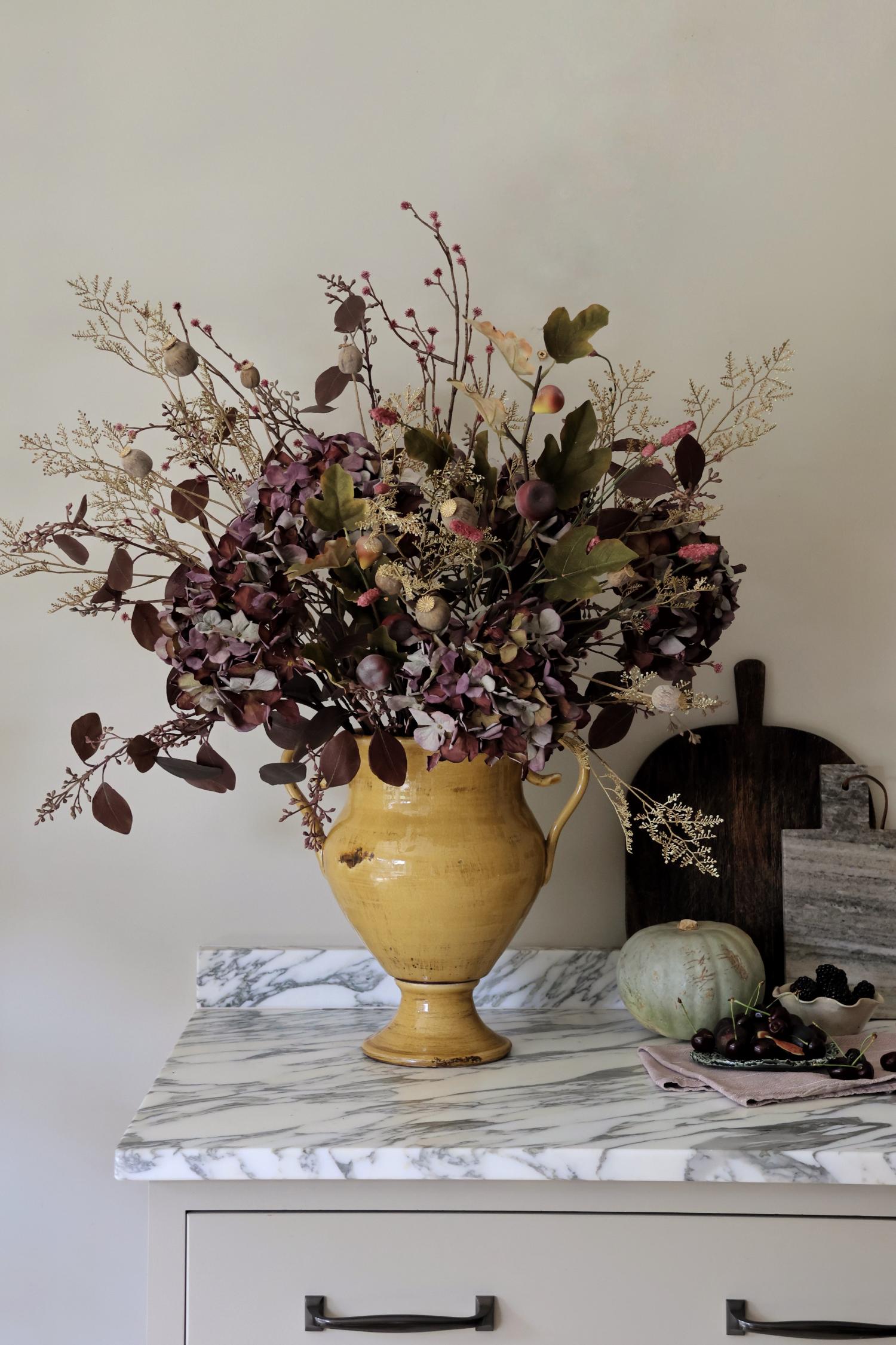 A large mustard ceramic vase filled with a artificial autumn floral arrangement in muted burgundy, beige, and olive tones, displayed on a white marble-topped cabinet against a light neutral wall.