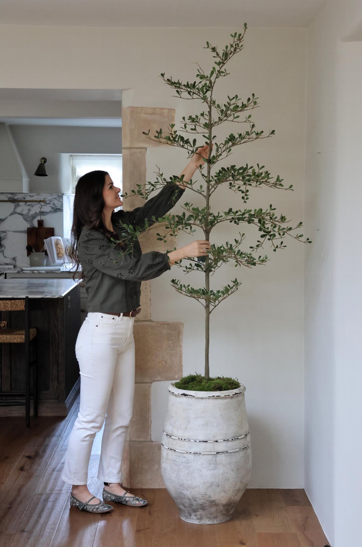 A person standing next to a tall, artificial black olive tree with green leaves, placed in a white pot.