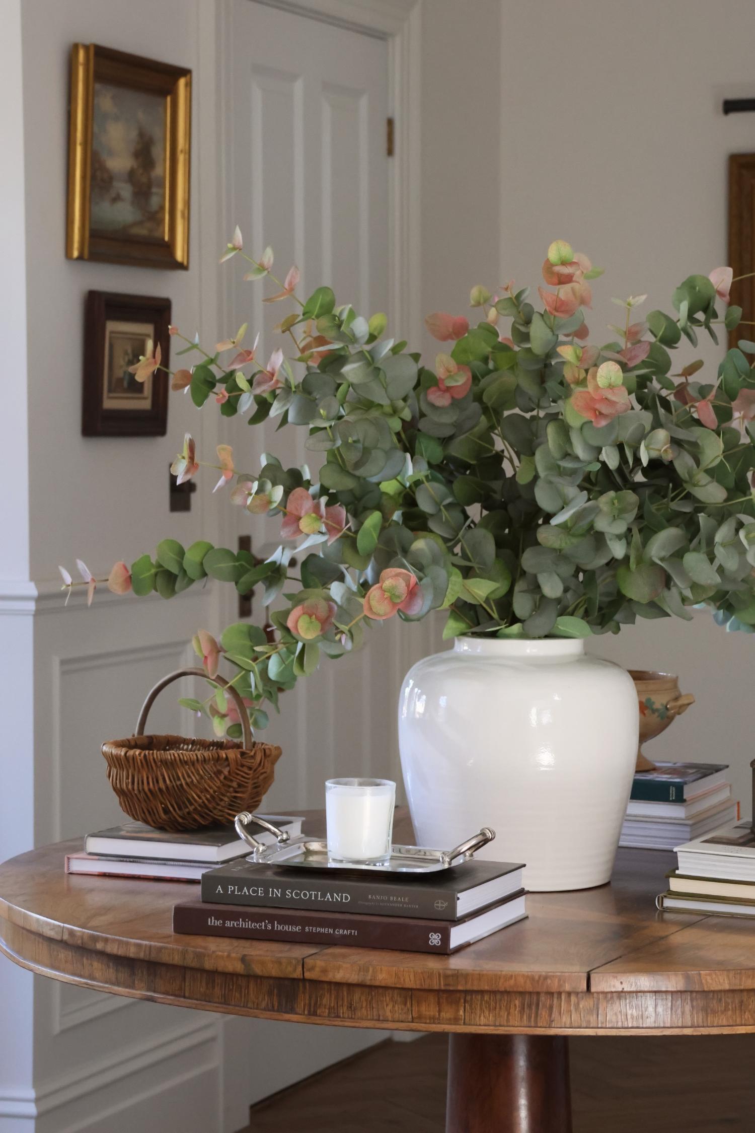 large white ceramic vase with artificial blush eucalyptus stems styled on a round wooden able with books and decor 
