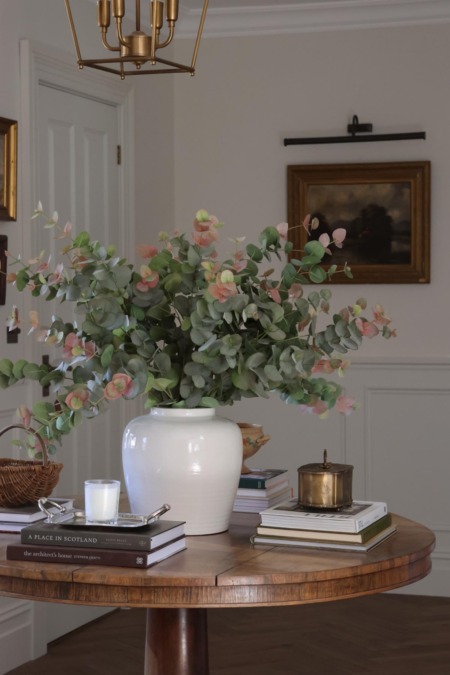 A large white ceramic vase filled with green eucalyptus stems and soft pink roses, placed on a round wooden table in a styled interior room with books, a candle, and wall art visible.