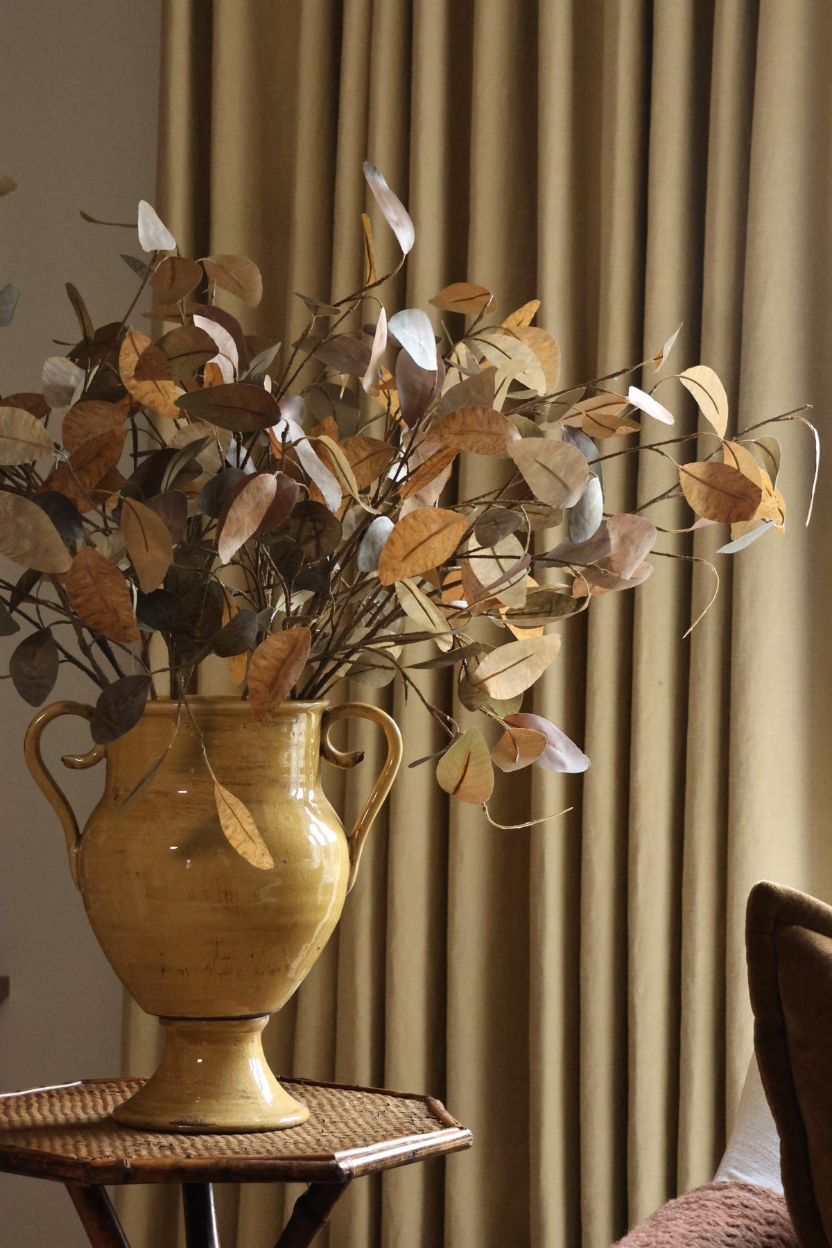 Artificial autumn foliage in large yellow urn with soft yellow curtains behind it 