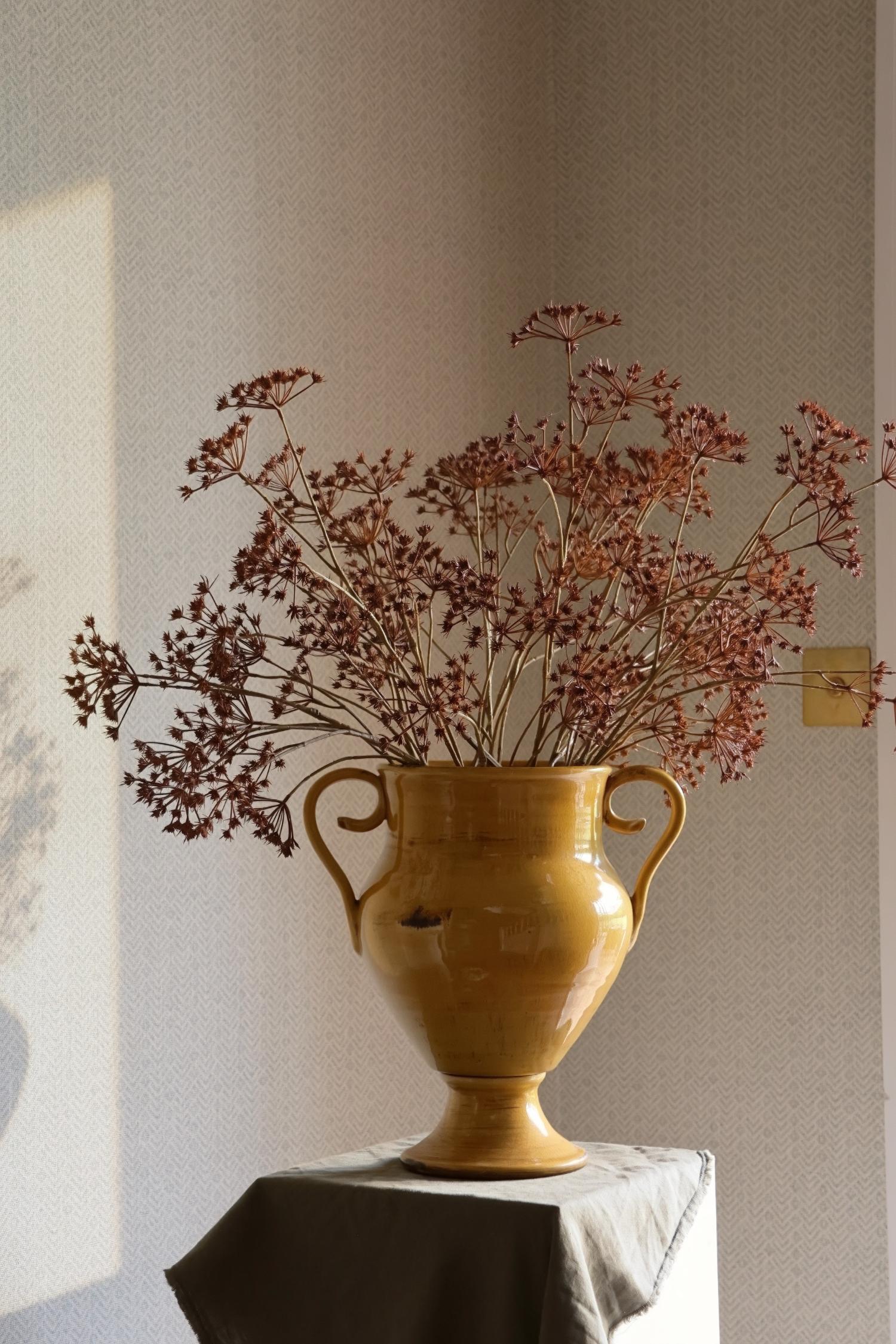Artificial autumn brown dried look stems in a yellow ceramic urn vase on pedestal table