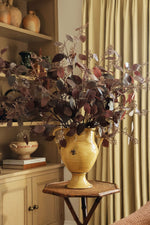 Artificial burgundy eucalyptus stems in a yellow urn vase with oak cabinetry and curtains behind 