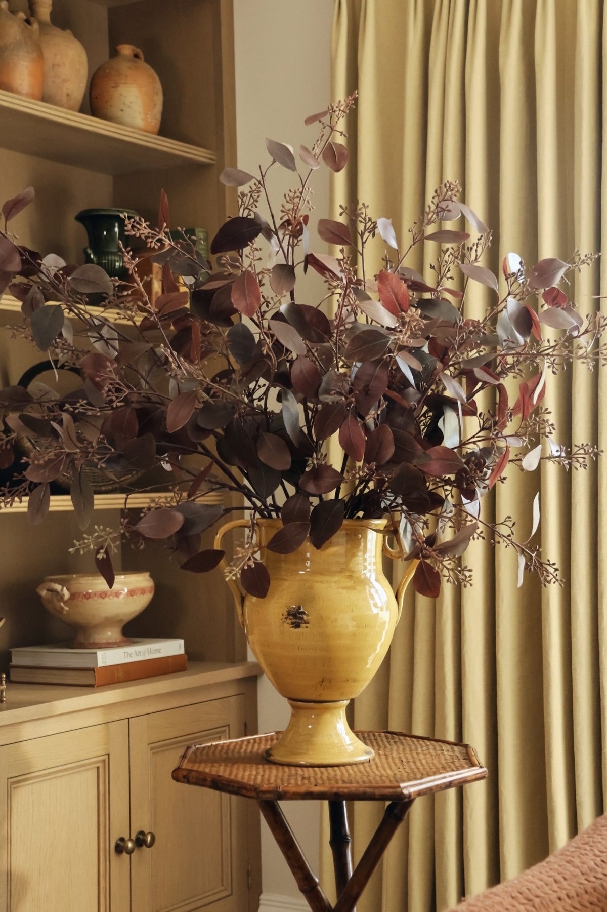 Artificial burgundy eucalyptus stems in a yellow urn vase with oak cabinetry and curtains behind 
