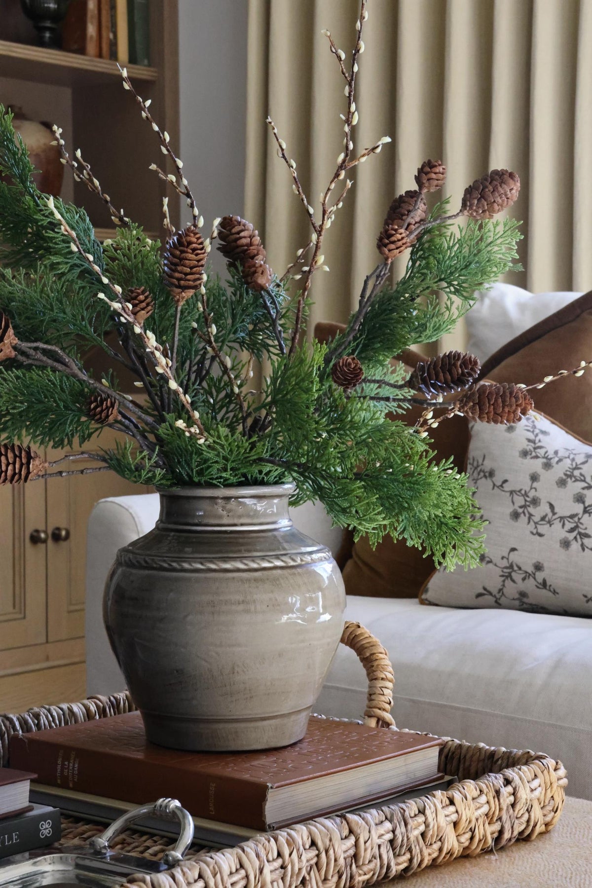 Artificial cypress branch and willow spray with cone branches styled in a grey ceramic vase on a coffee table in a cosy lounge setting 