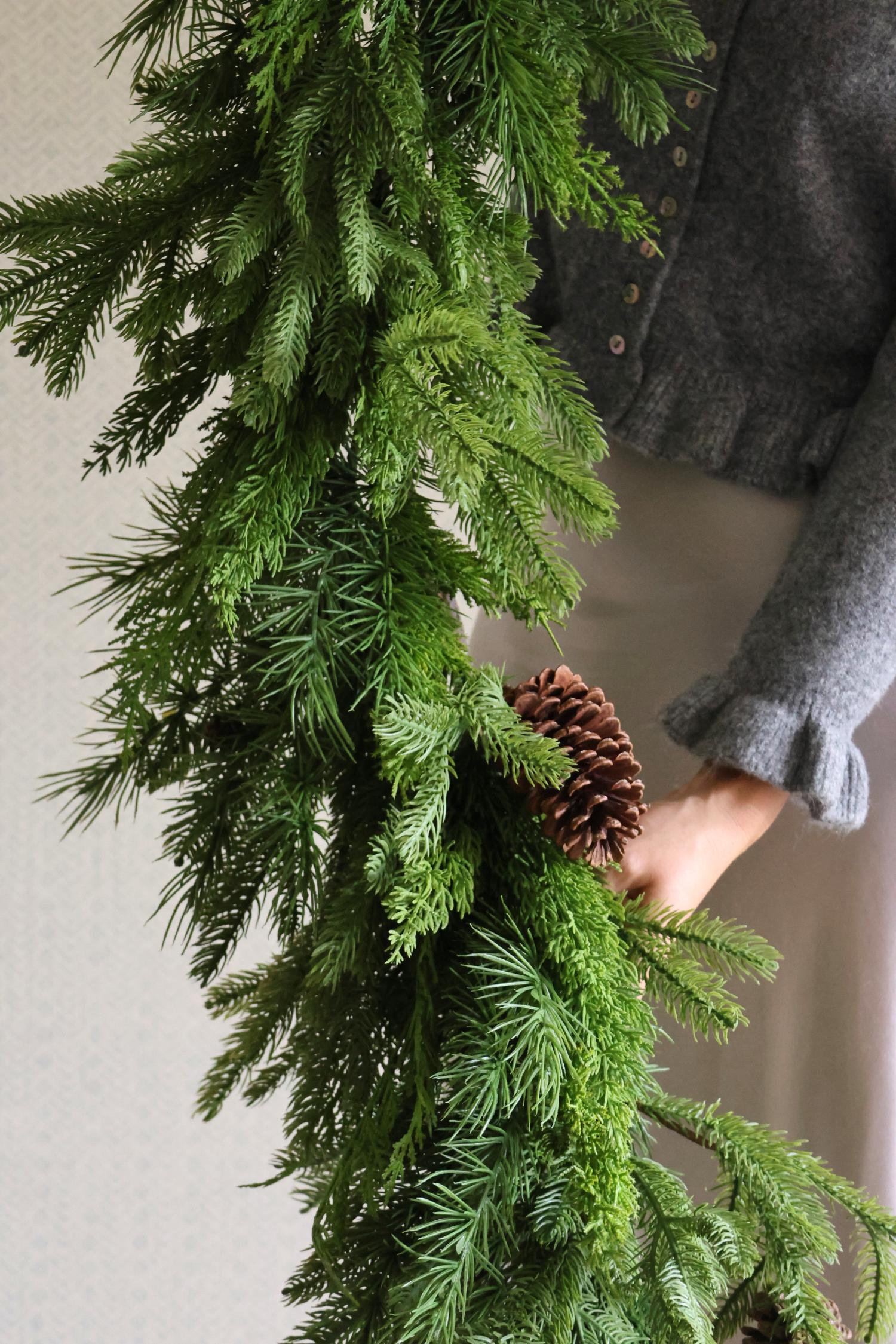 Person holding a large green artificial garland with a pine cone against a neutral background