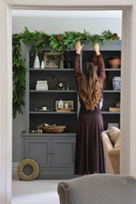 Person decorating a bookshelf with a large artificial mixed folIage garland  in a living room setting