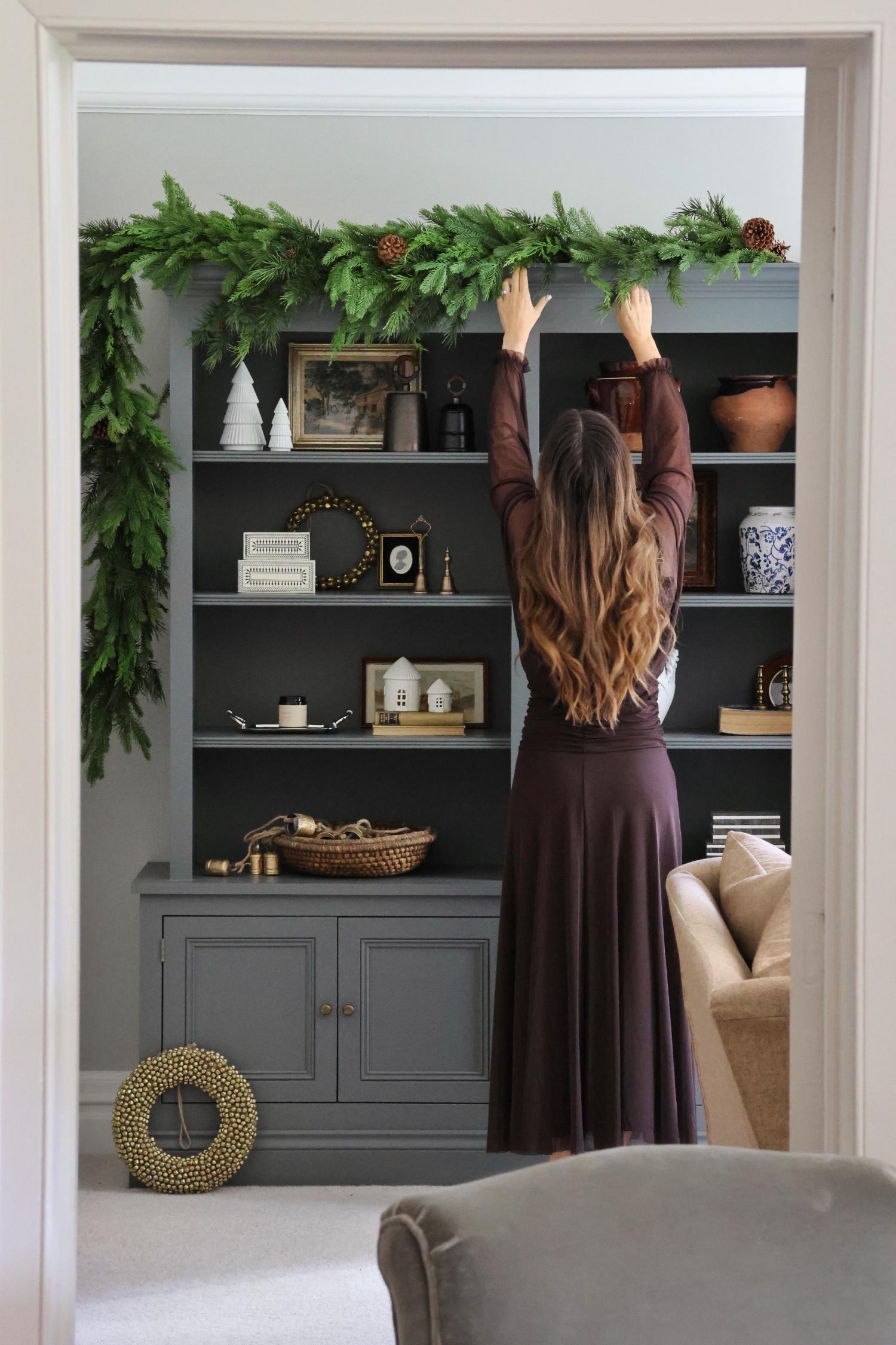 Person decorating a bookshelf with a large artificial mixed folIage garland  in a living room setting