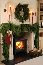 A cozy holiday fireplace scene with a green garland draped over a black wood-burning stove, a matching wreath on the mantel, red candles, a red Christmas stocking, and a basket of firewood on the floor.