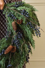 A close-up of an artificial green evergreen wreath decorated with dark blue berries, pinecones, and brown branches, held by a person wearing a brown plaid shirt against a beige wall.