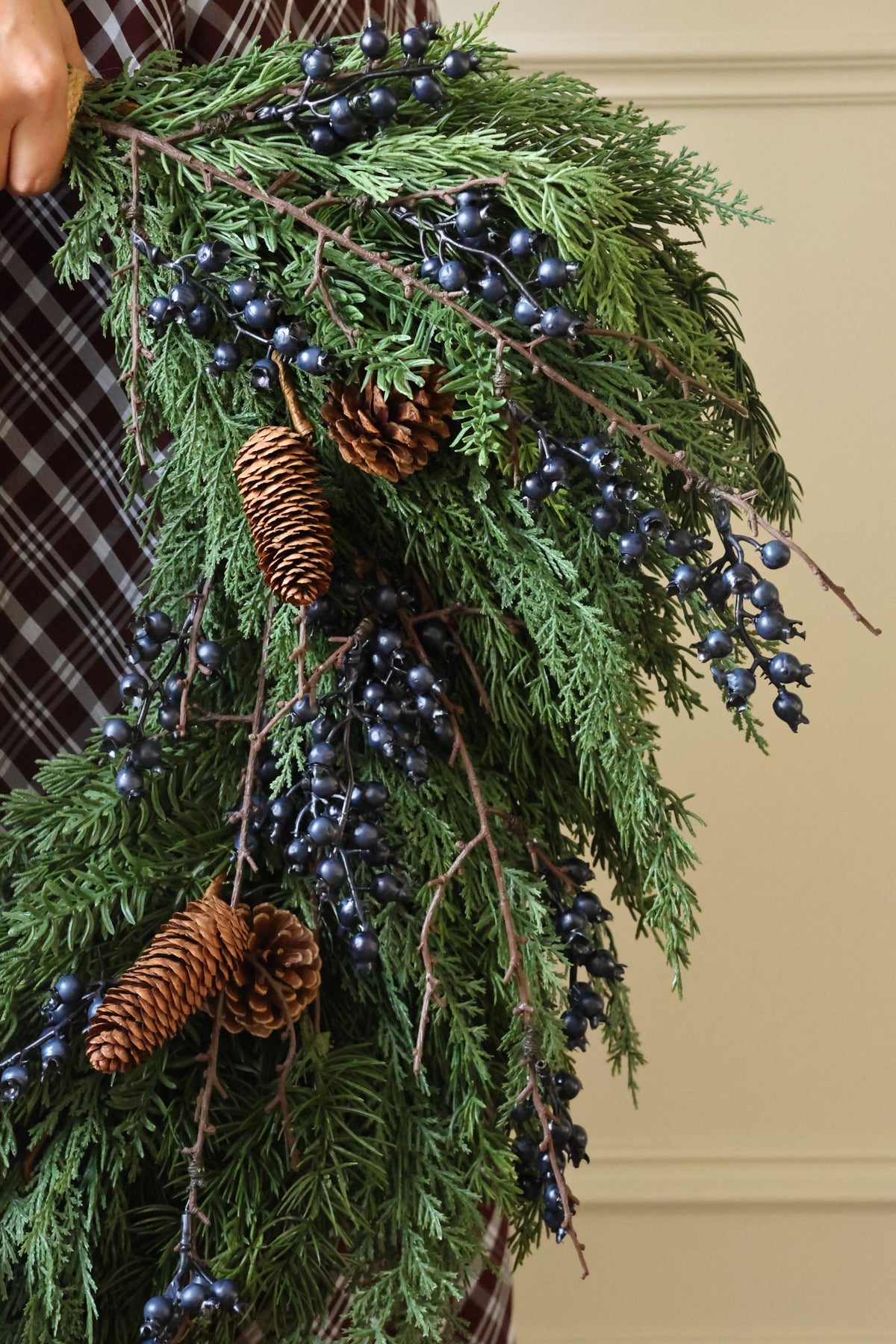 A close-up of an artificial green evergreen wreath decorated with dark blue berries, pinecones, and brown branches, held by a person wearing a brown plaid shirt against a beige wall.