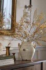 A large distressed ceramic vase filled with dried artificial autumn golden branches, displayed on a wooden console table in an elegant interior setting with a gold mirror and brass candlestick in the background.