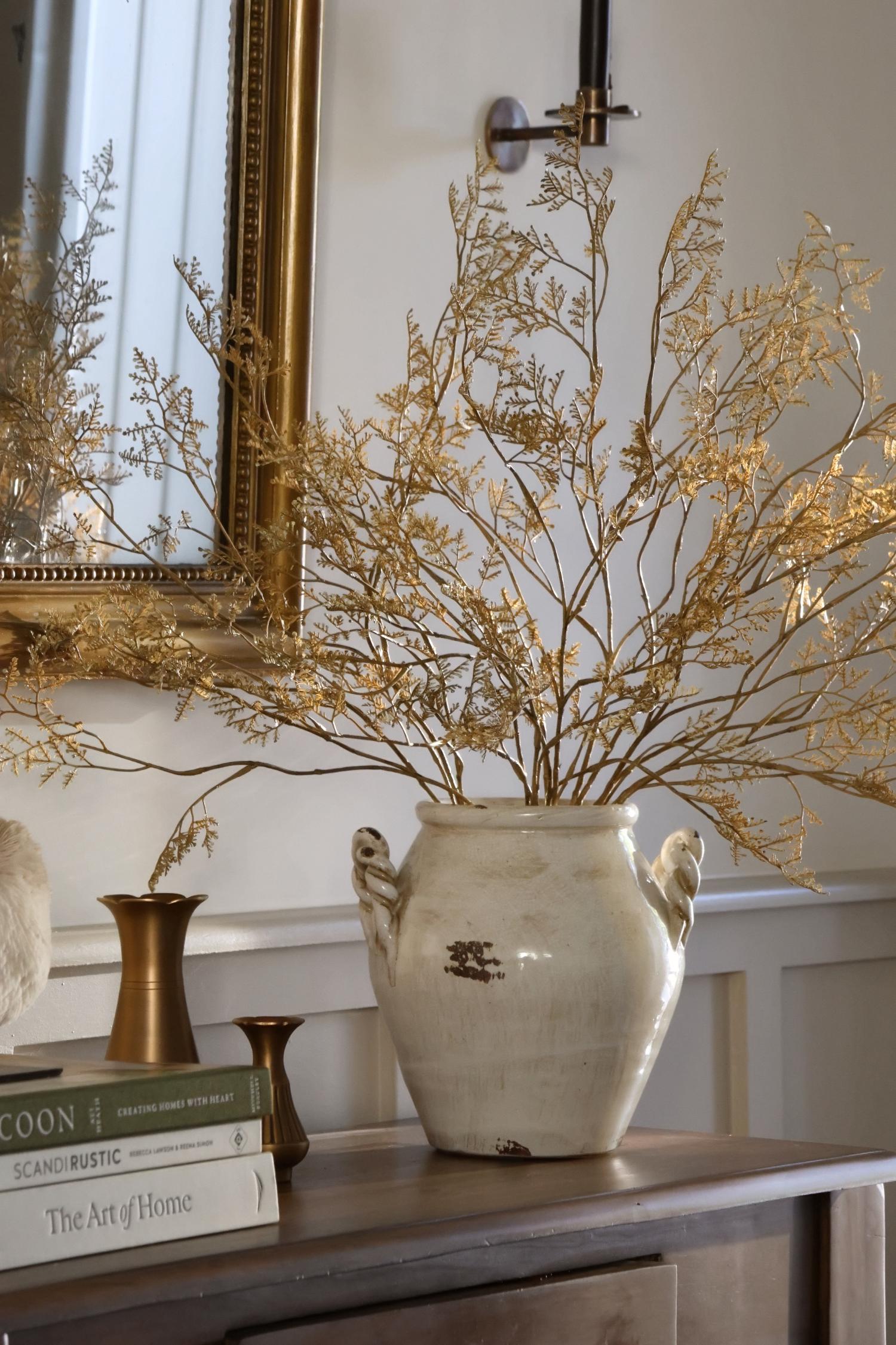 A large distressed ceramic vase filled with dried artificial autumn golden branches, displayed on a wooden console table in an elegant interior setting with a gold mirror and brass candlestick in the background.