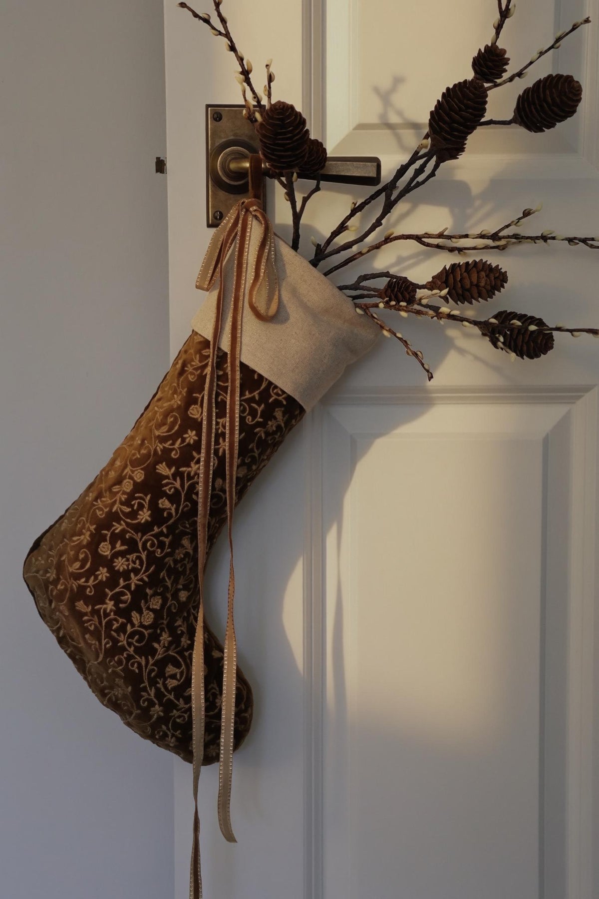 Embroidered bronze stocking hanging from white door with decorative branches inside 