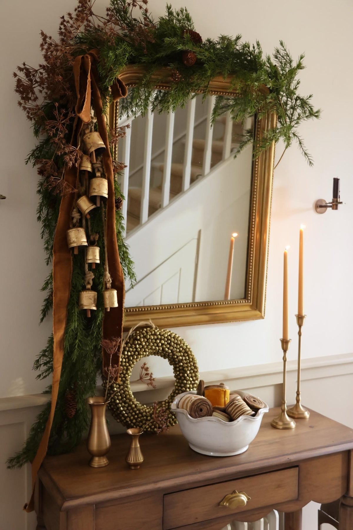 Christmas styled scene showing a gold mirror dressed with asymmetric green foliage garlands, brass cowbells, bronze ribbon and festive accessories on the console table including candlesticks and bud vases. 