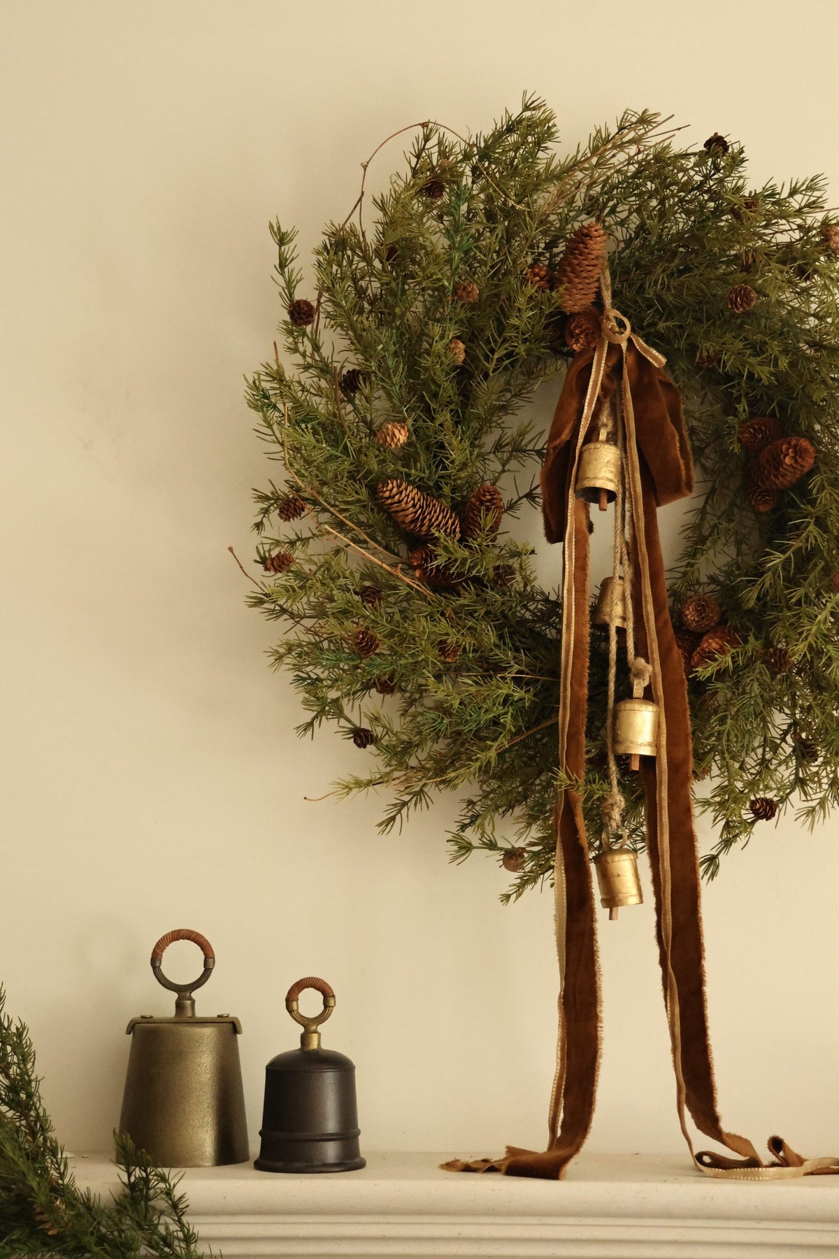 Large artificial foliage wreath with pinecones, brass cowbells, bronze ribbon on a fireplace