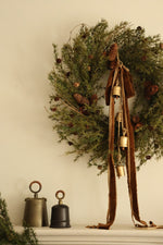 Large artificial foliage wreath with pinecones, brass cowbells, bronze ribbon on a fireplace