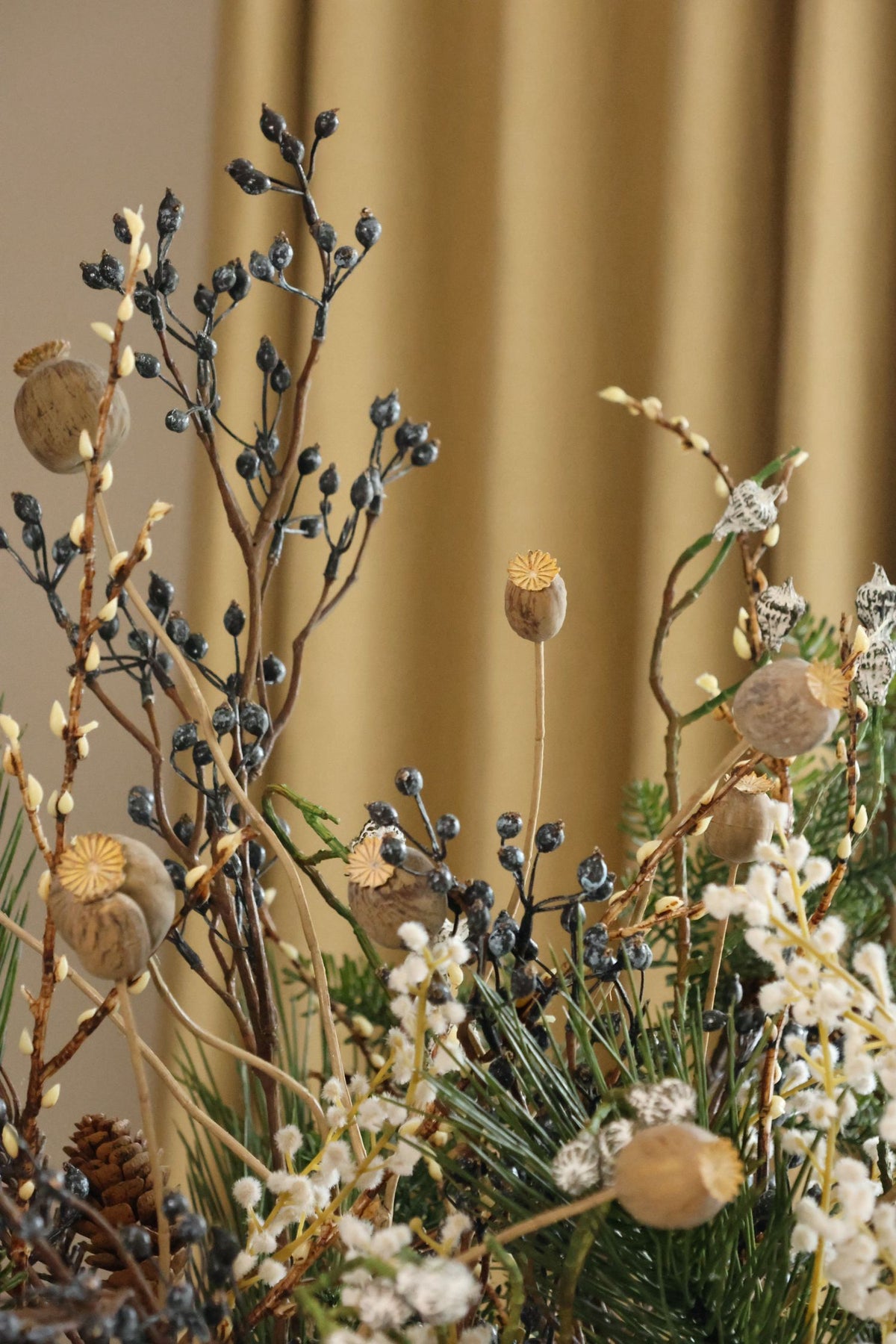 A close-up of an artificial floral arrangement featuring poppy pods, pinecones, white wildflowers, and dark blue berries against a warm beige curtain background.