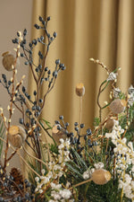 A close-up of an artificial floral arrangement featuring poppy pods, pinecones, white wildflowers, and dark blue berries against a warm beige curtain background.