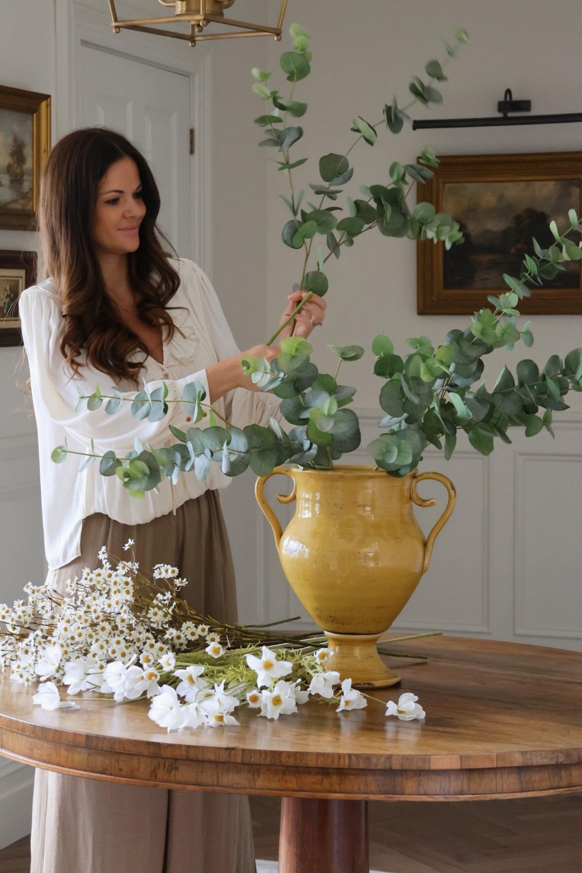 women arranging artificial green eucalyptus stems in a yellow urn on a wooden table 