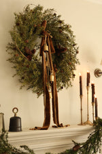 Faux nordic wreathwith velvet ribbons, cowbells and brass slim candlesticks ontop of a mantel 
