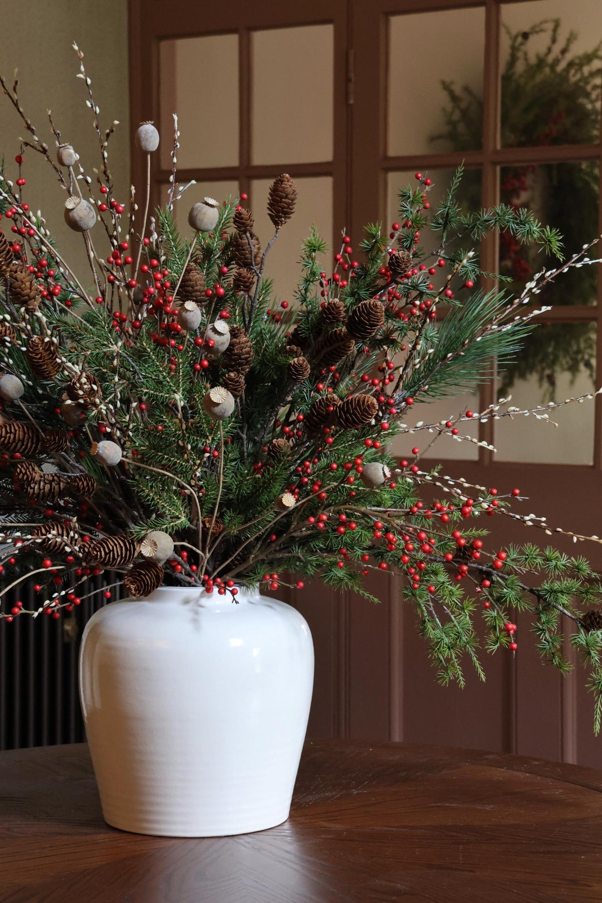 Decorative arrangement of greenery, red berries, and pinecones in a white vase on a wooden surface.